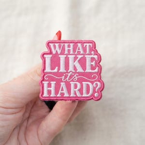 May include: A rectangular embroidered patch with a pink background and a scalloped border. The patch features white stitched text that reads "WHAT, LIKE it's HARD?" in a combination of bold block lettering and a cursive script font.