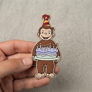 May include: An embroidered patch featuring a brown monkey wearing a red and yellow striped party hat. The monkey is holding a two-tiered birthday cake with light purple and light blue frosting, topped with seven lit yellow candles.
