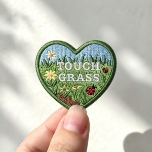 May include: A heart-shaped embroidered patch featuring a light blue sky, green grass, small white and yellow flowers, and two red ladybugs. The text "TOUCH GRASS" is stitched in white capital letters across the center of the patch.