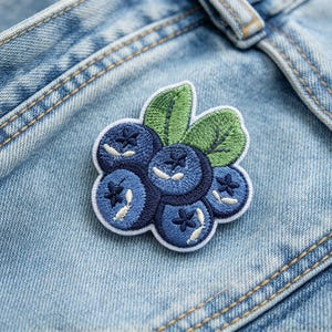 May include: An embroidered patch featuring a cluster of five round blueberries in shades of dark and medium blue, accented with two green leaves. The patch has a white border and is attached to light blue denim fabric with visible stitching.