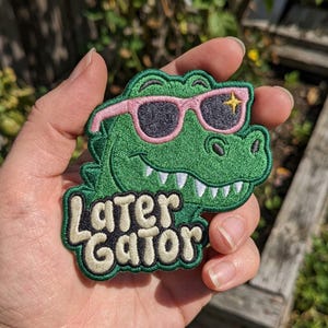 May include: An embroidered patch featuring a green alligator wearing pink sunglasses with a yellow star accent. The patch includes the text "Later Gator" in cream-colored, rounded lettering with a black outline, set against a dark green background.