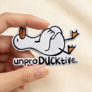 May include: An embroidered iron-on patch featuring a white duck with an orange beak and feet, lying on its back with its wings tucked in. Below the duck, the black embroidered text reads unproDUCKtive. The patch has a white border.
