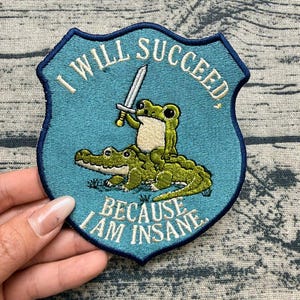 May include: A shield-shaped embroidered patch with a teal background and a dark blue border. It features a green frog riding a green crocodile while holding a silver sword. The text reads I WILL SUCCEED BECAUSE I AM INSANE in white lettering.