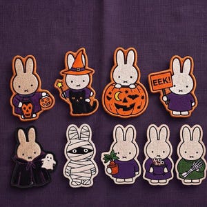 May include: A set of eight embroidered patches featuring white rabbit characters in various Halloween costumes, including a witch, a mummy, a vampire, and a pumpkin. The patches display vibrant orange, purple, and black thread details.
