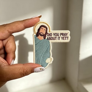 May include: An embroidered patch featuring a stylized figure with brown hair and a beard, wearing a teal robe with a gold halo. A speech bubble extends to the right with the dark brown embroidered text: DID YOU PRAY ABOUT IT YET?