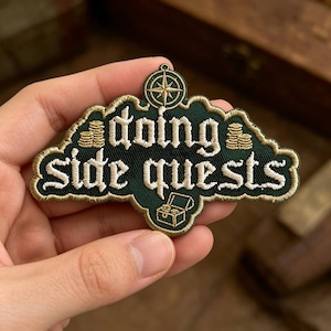 May include: An embroidered patch with a dark green background and gold border. It features the text "doing side quests" in white, a gold compass icon at the top, two stacks of gold coins, and an open treasure chest icon at the bottom.