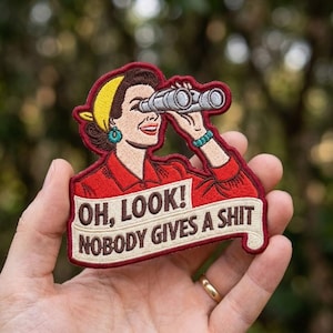 May include: An embroidered patch featuring a retro-style illustration of a person wearing a yellow headscarf and red shirt, looking through grey binoculars. A cream-colored banner across the bottom displays the text: OH, LOOK! NOBODY GIVES A SHIT.
