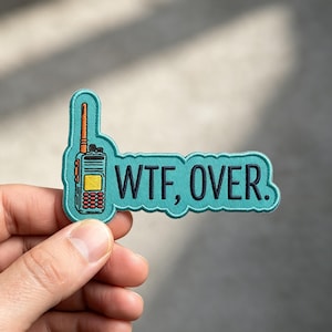 May include: A teal embroidered patch featuring a graphic of a two-way radio with an orange antenna and yellow screen, accompanied by the black embroidered text "WTF, OVER." The patch has a die-cut shape with a matching teal stitched border.