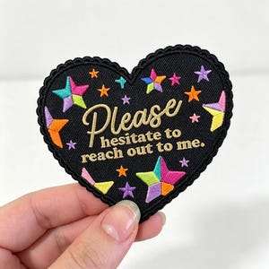 May include: A heart-shaped black embroidered patch with a scalloped edge. The patch features gold text that reads "Please hesitate to reach out to me." and is decorated with various multi-colored embroidered stars in shades of pink, blue, and orange.