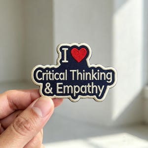 May include: A dark blue embroidered patch with a cream-colored border. The text reads I (with a red heart symbol) Critical Thinking & Empathy in cream-colored lettering. The patch has a scalloped, irregular shape following the text outline.