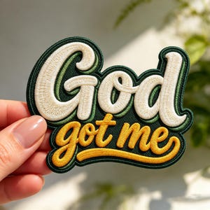 May include: An embroidered patch featuring the text God in cream-colored thread and the text got me in yellow thread. The lettering is set against a dark green background with a thick dark green border outlining the entire shape.