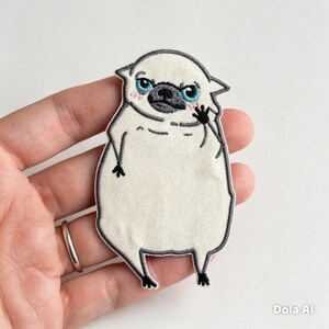 May include: An embroidered patch featuring a stylized, round, cream-colored creature with a dark gray snout, bright blue eyes, and pink cheeks. The figure has small black limbs, a dark gray border, and the text Dola AI in the bottom right corner.