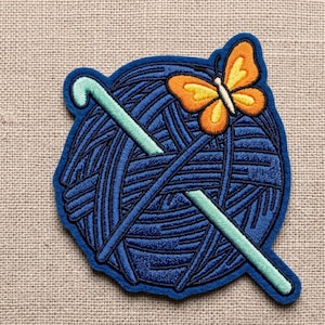 May include: Embroidered patch featuring a blue yarn ball with two crossed crochet hooks in light green. An orange and yellow butterfly sits atop the yarn ball. The patch has a dark blue border and is on a tan fabric background.