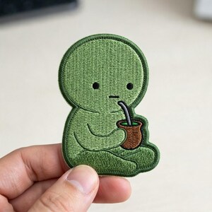 May include: An embroidered patch featuring a green, rounded character sitting in a cross-legged position. The character has two small black eyes, a straight line for a mouth, and is holding a brown mate gourd with a silver straw in its hands.