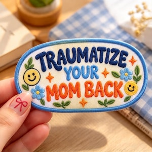 May include: An oval-shaped embroidered patch with a cream-colored background and a blue border. The text reads "TRAUMATIZE YOUR MOM BACK" in blue and orange letters. The design includes yellow smiley faces, blue flowers, green leaves, and orange stars.