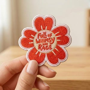 May include: A flower-shaped embroidered patch with red petals, a light pink border, and a cream-colored center. The text "FULL of WHIMSY AND RAGE" is stitched in red in the center of the patch, surrounded by small radiating lines.