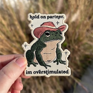 May include: An embroidered patch featuring a green frog wearing a pink cowboy hat. The text above the frog reads hold on partner, and the text below reads im overstimulated. The design includes small black star accents on a cream background.
