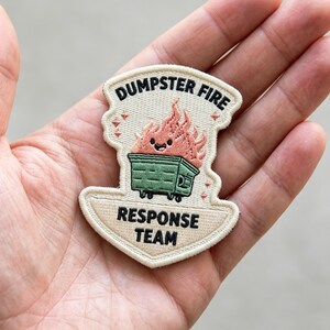 May include: An embroidered patch with a cream-colored border featuring a green dumpster with a smiling pink flame character on top. The text DUMPSTER FIRE is stitched in black at the top, and RESPONSE TEAM is stitched in black at the bottom.