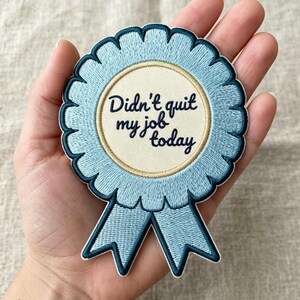 May include: A circular embroidered patch shaped like a blue ribbon award. The center is cream-colored with the dark blue text "Didn't quit my job today" written in a cursive font. The patch features a scalloped edge and two downward-pointing tails.