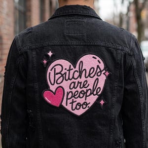 May include: A black denim jacket featuring a large embroidered patch on the back. The patch is a pink heart shape with the black text "Bitches are people too." It includes a smaller dark pink heart and several small black star-shaped accents.