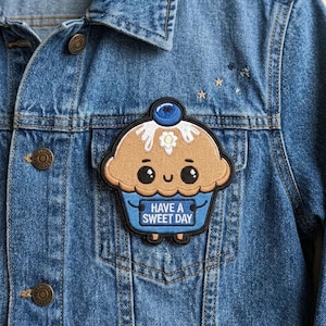May include: A blue denim jacket featuring an embroidered patch of a tan cupcake with a blue wrapper, a blueberry on top, and a white flower. The cupcake has a smiling face and holds a blue sign with the white text HAVE A SWEET DAY.