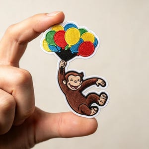 May include: An embroidered patch featuring a brown monkey holding a cluster of colorful balloons in red, yellow, blue, and green. The monkey has a smiling expression and is suspended by black strings attached to the vibrant balloon bunch.