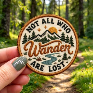 May include: A circular embroidered patch with a cream background and a brown border. It features a mountain range, a yellow sun, green pine trees, and a blue river. The text reads NOT ALL WHO WANDER ARE LOST in black and brown.