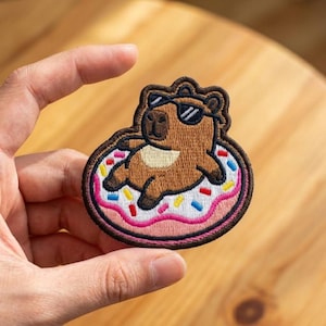May include: An embroidered patch featuring a brown capybara wearing black sunglasses while floating on a white donut-shaped pool float with pink icing and colorful sprinkles. The patch has a dark brown stitched border and a detailed thread texture.