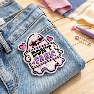 May include: An embroidered iron-on patch featuring a light pink ghost wearing black star-shaped sunglasses. The ghost holds a rectangular sign with the black and purple text DON'T PANIC, surrounded by three small pink hearts on a blue denim fabric.