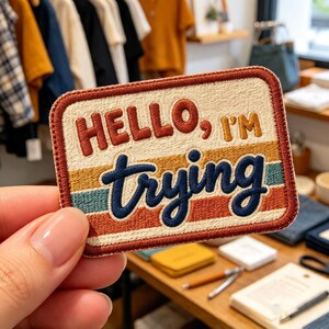 May include: A rectangular embroidered patch with a cream-colored background and a rust-colored border. The text reads HELLO, I'M in rust and gold, with the word trying in dark blue cursive script over horizontal stripes of rust, gold, and teal.