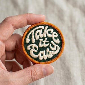 May include: A circular embroidered patch featuring a dark green background with the text Take it easy stitched in cream-colored thread. The patch is finished with a vibrant orange border that frames the central design in a clean, circular shape.