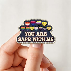 May include: A dark blue embroidered patch with a scalloped edge featuring the text "YOU ARE SAFE WITH ME" in cream-colored letters. Above the text are eleven small hearts, each filled with different pride flag color patterns in various stripes.