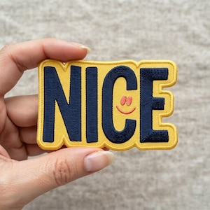 May include: A rectangular embroidered patch with a yellow background and a dark blue border. The word NICE is stitched in dark blue capital letters. The letter C features a small, orange, smiling face graphic centered within its curve.