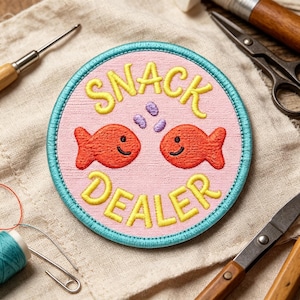 May include: A circular embroidered patch with a light pink background and a bright blue border. The patch features two orange fish facing each other, three small purple dots, and the yellow embroidered text SNACK DEALER.