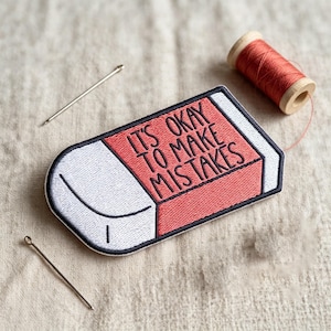 May include: An embroidered patch shaped like a rectangular eraser with a white end and a coral-colored end featuring the black embroidered text IT'S OKAY TO MAKE MISTAKES, accompanied by two silver sewing needles and a spool of coral thread.