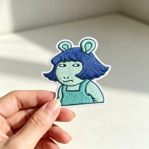 May include: An embroidered patch featuring a cartoon character with a light green face, dark blue hair, and a matching dark blue top. The character has a skeptical expression with narrow eyes and a small mouth, outlined in white thread on a fabric base.