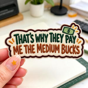 May include: An embroidered patch with a cream-colored background and a dark brown scalloped border. It features the green text THAT'S WHY THEY PAY and brown text ME THE MEDIUM BUCKS, with a small green dollar bill icon in the top right corner.