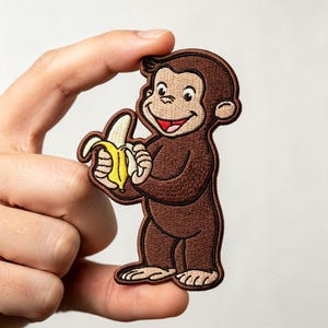 May include: An embroidered iron-on patch featuring a brown monkey with a tan face, ears, hands, and feet, holding a yellow banana. The monkey has a smiling expression with a visible red tongue and is outlined in dark brown thread on a white background.