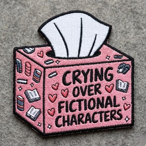 May include: A pink embroidered patch shaped like a tissue box with a white tissue emerging from the top. The box features black text reading CRYING OVER FICTIONAL CHARACTERS, surrounded by small red hearts, open books, and dark blue book stacks.