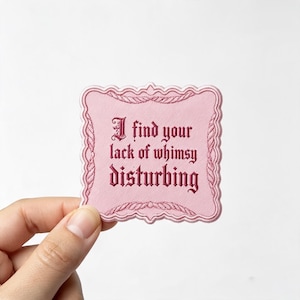 May include: A square embroidered patch with a light pink background and a decorative scalloped border. The text "I find your lack of whimsy disturbing" is stitched in a dark red gothic-style font across the center of the patch.