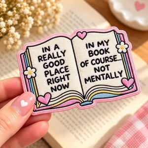 May include: An embroidered patch shaped like an open book with pink borders. The left page reads IN A REALLY GOOD PLACE RIGHT NOW and the right page reads IN MY BOOK OF COURSE... NOT MENTALLY. It features small pink hearts and white flowers.