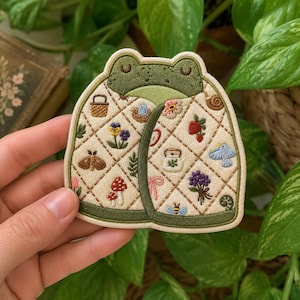 May include: A cream-colored embroidered patch shaped like a frog head, featuring a quilted diamond pattern. The design includes small motifs of a basket, flowers, a strawberry, a moth, mushrooms, a bird, a bee, and a teapot, all bordered by dark green trim.