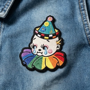 May include: An embroidered patch featuring a crying baby clown with a rainbow-colored ruffled collar. The clown wears a teal party hat decorated with three small flowers and a black and white checkered brim, all outlined in thick black thread.