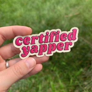May include: An embroidered patch with a cream-colored background and a dark pink border. The text "certified yapper" is stitched in dark pink, bold, rounded letters across the center of the patch, which has a wavy, irregular shape.