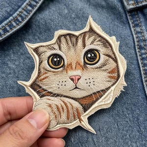 May include: An embroidered patch featuring a brown tabby cat with large golden eyes and a pink nose, depicted as if peeking through a torn opening. The design includes detailed stitching of fur patterns, whiskers, and a cream-colored border.
