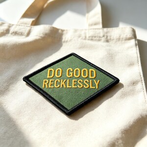 May include: A natural-colored canvas tote bag featuring a diamond-shaped patch. The patch has a dark green background with a thick black border and the yellow embroidered text "DO GOOD RECKLESSLY" centered in the middle of the fabric surface.