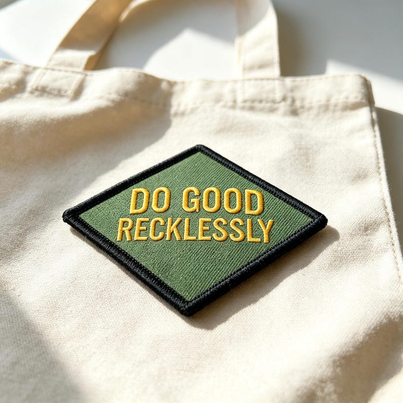May include: A natural-colored canvas tote bag featuring a diamond-shaped patch. The patch has a dark green background with a thick black border and the yellow embroidered text "DO GOOD RECKLESSLY" centered in the middle of the fabric surface.