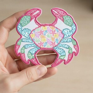 May include: An embroidered crab-shaped patch featuring a white background with a colorful pattern of seashells and starfish. The crab has pink claws, light blue and green legs with dark blue spots, and a bright pink stitched border outline.