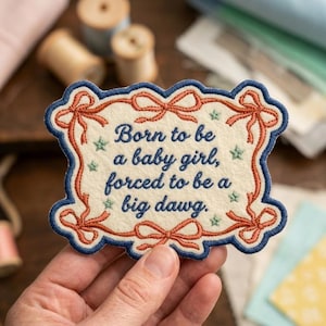 May include: An off-white embroidered patch with a dark blue border and orange ribbon motifs at the corners. The text reads: Born to be a baby girl, forced to be a big dawg. Small green stars are scattered around the dark blue embroidered lettering.