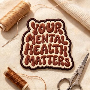 May include: An embroidered patch with a scalloped edge featuring the dark brown text YOUR MENTAL HEALTH MATTERS on a cream background. The patch is surrounded by two spools of brown thread and a pair of silver metal sewing scissors.
